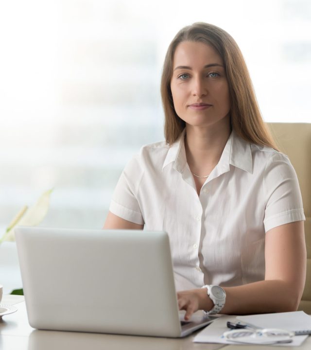 Female office worker doing daily work on laptop Portrait of confident businesswoman sitting at desk in office and working on laptop. Attractive woman typing on computer and looking at camera. Female office worker doing daily work with pleasure